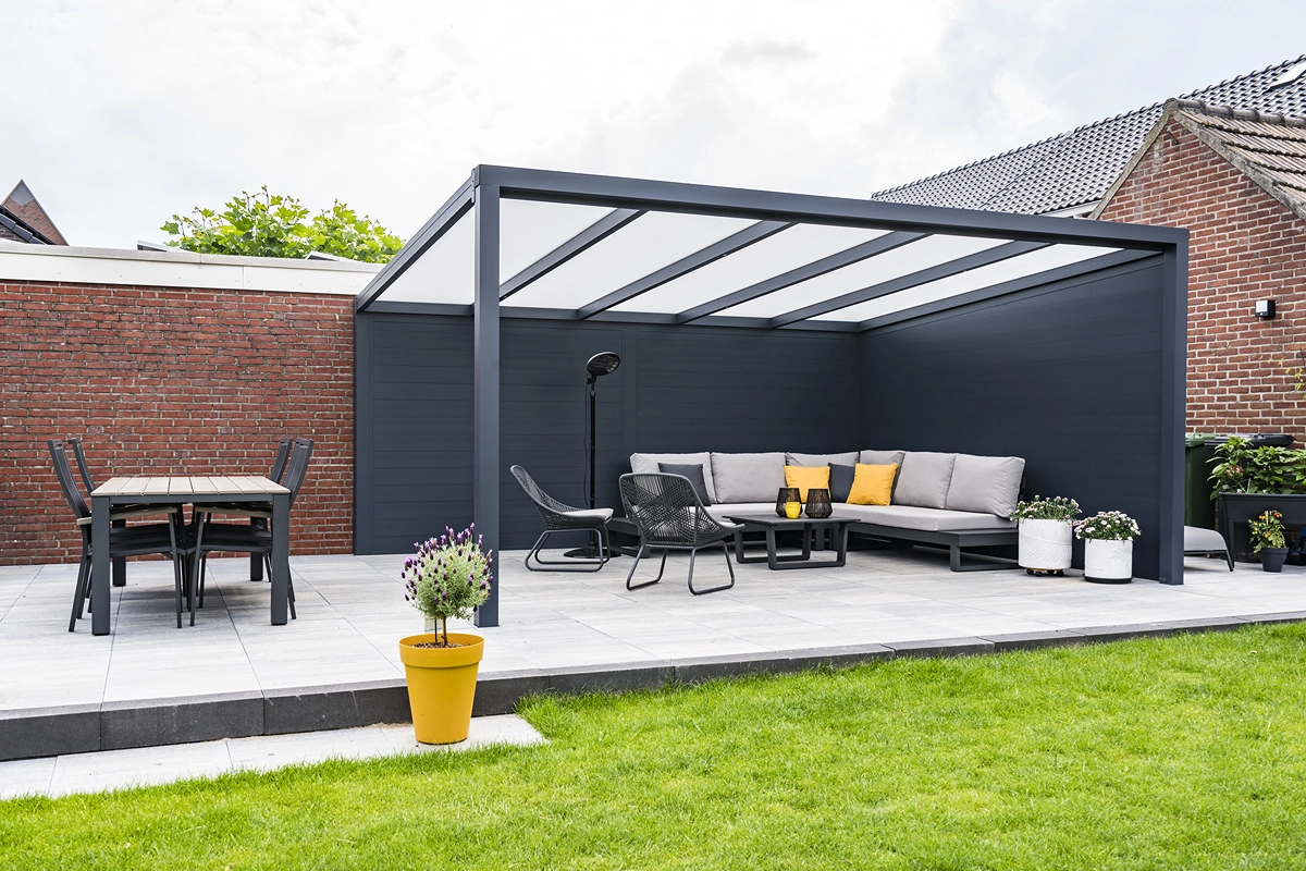 Moderne anthrazitfarbene Terrassenüberdachung mit Lounge-Sofa, Esstisch, Pflanzen und gelbem Pflanzkübel auf gepflasterter Terrasse